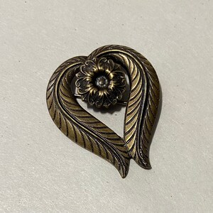 May include: A gold-toned heart-shaped brooch with a detailed floral design. The center of the flower is a circular shape with a raised center.