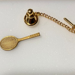 May include: A gold-toned tie tack featuring a tennis racket and a chain with a clasp.
