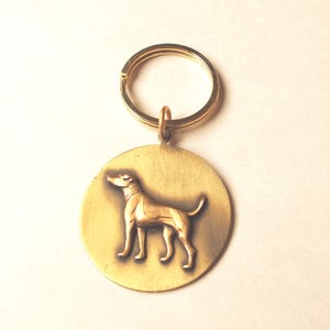 May include: A circular brass keychain featuring a raised, three-dimensional relief of a standing dog in profile. The metal has a brushed, matte gold-toned finish, and the design is attached to a standard round split ring for keys.