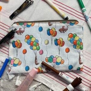 May include: A white zippered pouch with a repeating pattern of monkeys holding colorful balloons. The pouch is on a white and pink striped towel with other items, including a nail file, a pink lip gloss, and a brown marker.