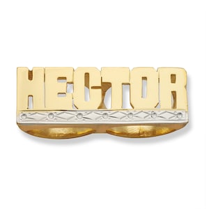 May include: A gold-toned ring with the name "HECTOR" in block letters. The ring features a two-finger design with a decorative band of silver-toned metal and small, clear accents. The ring is likely made of gold-plated metal.