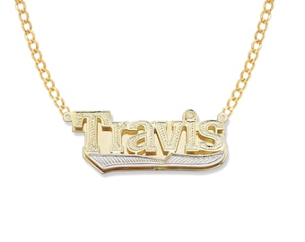 May include: A gold-colored necklace with the name "Travis" in a layered, block-letter style. The pendant is two-toned, with a gold top layer and a silver-colored base. The chain is a delicate gold link chain.