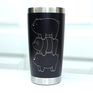 May include: A black stainless steel tumbler featuring a laser-engraved white line art design of three cartoon bears stacked vertically on top of each other. The cup has a metallic silver rim at the top and a matching metallic silver base.