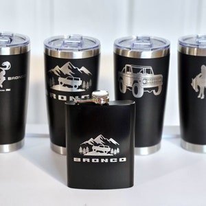 May include: Set of four black stainless steel tumblers and a black stainless steel flask, all with silver engraved designs featuring a Bronco logo and various Bronco-themed images, including a horse, mountains, and a truck.