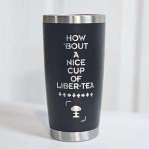May include: A dark navy blue stainless steel tumbler with a silver rim and base. The front features the engraved text "HOW 'BOUT A NICE CUP OF LIBER-TEA" above a row of directional arrows and a small mushroom cloud icon centered in brackets.