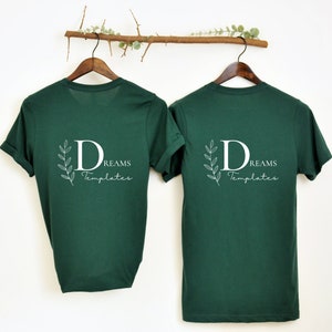 May include: Two dark green t-shirts with short sleeves hanging on wooden hangers. The t-shirts have a white graphic on the back that says "DREAMS Templates" with a stylized letter "D" and leaves.