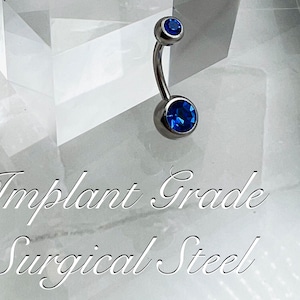 May include: A surgical steel belly button ring with two blue gem stones.  The ring is on a white background with the text "Implant Grade Surgical Steel".