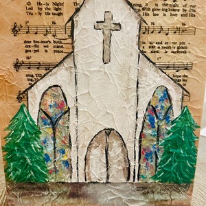 Puede incluir: Una obra de arte navide&ntilde;a hecha a mano que representa una iglesia blanca con una cruz, vidrieras y &aacute;rboles de Navidad verdes. El fondo es una partitura musical con el texto "O Holy Night". La obra est&aacute; sobre una superficie texturizada.