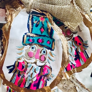 May include: A hand-painted oyster shell ornament featuring a whimsical character with a pink face, blue eyes, and a teal hat. The character is wearing a pink and gold striped jacket and has a white beard. The ornament is decorated with gold accents and is perfect for adding a touch of whimsy to your holiday decor.