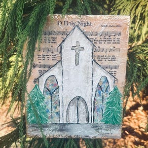 Puede incluir: Una obra de arte texturizada hecha a mano que representa una iglesia blanca con una cruz, vidrieras y &aacute;rboles verdes. El fondo es una partitura musical con las palabras "O Holy Night".
