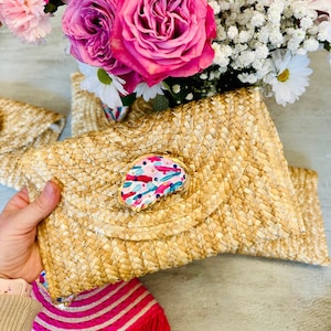 Puede incluir: Un bolso de mano de paja con un diseño de concha pintada de colores. El bolso está cerrado y tiene una solapa.