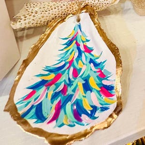 Puede incluir: Adorno navideño de concha de ostra pintado a mano. La concha blanca presenta un diseño de árbol de Navidad colorido con pinceladas azules, rosas, amarillas y verdes. La concha tiene un borde dorado y está atada con una cinta dorada.