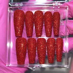 Puede incluir: Un conjunto de diez u&ntilde;as postizas de color rojo con purpurina en una caja de pl&aacute;stico transparente. Las u&ntilde;as son largas y de forma de ata&uacute;d.