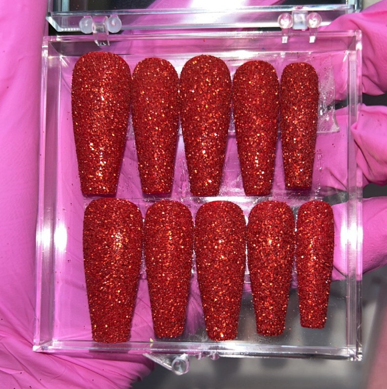 Puede incluir: Un conjunto de diez u&ntilde;as postizas de color rojo con purpurina en una caja de pl&aacute;stico transparente. Las u&ntilde;as son largas y de forma de ata&uacute;d.
