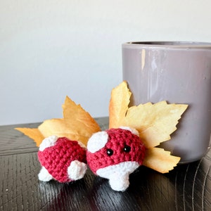 May include: Two small, handmade crochet mushrooms with red caps and white spots and stems. The mushrooms are placed near a gray vase and a dried leaf on a dark wooden surface. The mushrooms have black eyes.
