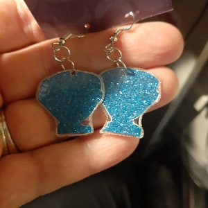 Op de afbeelding: Een paar blauwe glitter oorbellen in de vorm van schelpen. De oorbellen zijn aan een zilveren haak.