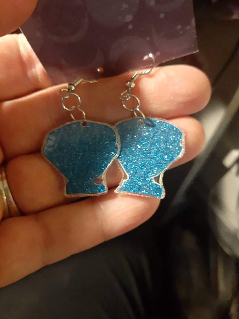 Op de afbeelding: Een paar blauwe glitter oorbellen in de vorm van schelpen. De oorbellen zijn aan een zilveren haak.