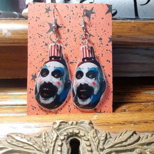 May include: A pair of dangle earrings featuring a horror-themed design. Each earring showcases a face with white, blue, and red accents, with a red and white striped hat. The earrings are suspended from silver-colored hooks and are set against an orange background.