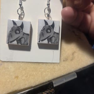 May include: A pair of dangle earrings featuring square charms with a black and white illustration of a dog with large eyes and pointed ears. The charms are attached to silver-toned metal hooks and are displayed on a white rectangular card.