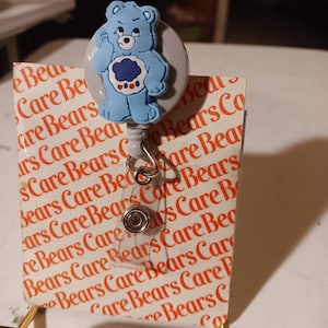 May include: A blue Care Bear character with a cloud on its belly is featured on a retractable badge holder. The holder is attached to a white background with the words "Care Bears" repeated in orange.