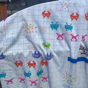 May include: White fabric with a grid pattern and colorful pixelated alien graphics. The design includes red, green, blue, and pink creatures, along with starbursts. The fabric has a blue border and text that reads "Space Invaders".