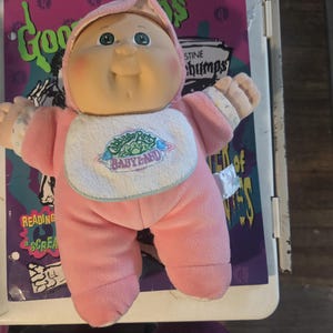 May include: A Cabbage Patch Kids doll with a soft vinyl head, green eyes, and rosy cheeks. The doll wears a pink fleece onesie with a matching hood and a white bib featuring the embroidered text Cabbage Patch Kids BABYLAND in green and pink.
