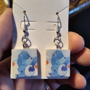 May include: A pair of blue Care Bear earrings with silver hooks. The earrings are square and feature a blue Care Bear with a white belly and a blue cloud on its belly. The Care Bear is holding a heart in its paws.