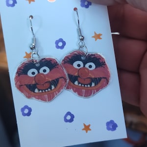 May include: A pair of dangle earrings featuring a red, furry character with a toothy grin. The character's eyes are wide and black, and the earrings are made of clear acrylic.