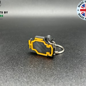 May include: Black and orange engine light keyring with a silver key ring. The keyring is made of plastic and has a textured surface.