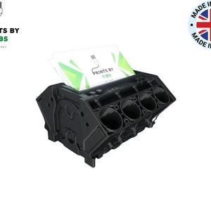 May include: Black plastic business card holder shaped like a car engine. The holder has a slot for business cards and the text "PRINTS BY TIBS" is printed on the business cards.