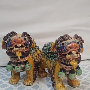 Puede incluir: Dos figuras de leones guardianes de cerámica con cuerpos amarillos, melenas marrones y detalles en verde azulado. Cada león tiene la boca abierta con dientes visibles y un collar con tres campanas verdes colgantes. Una cinta mide 25,4 cm.