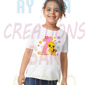Puede incluir: Una camiseta blanca con un gráfico colorido para un primer cumpleaños. El diseño incluye un gran número rosa "1" con una abeja de dibujos animados, flores y el texto "AY CREATIONS SAN ANTONIO". El niño lleva vaqueros azules.