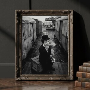 May include: A framed black and white photograph of a person in a bowler hat smoking a cigarette, set against a backdrop of trailers. The photograph is in a decorative brown frame, resting on a wooden floor. A stack of books and a lit candle are nearby.