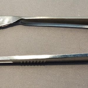 Puede incluir: Una pinza para hielo plateada de época. La pinza tiene una cabeza plana en forma de pala y un mango curvo. El mango tiene un agarre estriado. El metal muestra signos de desgaste. La herramienta es probablemente de acero inoxidable.