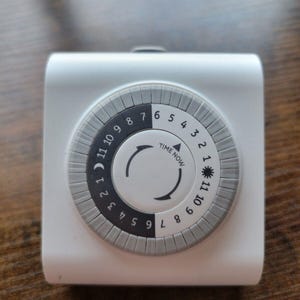 May include: A white, square-shaped mechanical timer with a grey dial. The dial features numbers 1-12, a sun and moon symbol, and the words "TIME NOW". The timer is designed for controlling electrical devices.
