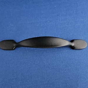 Może przedstawiać: Czarny uchwyt do szafki z zakrzywionym środkiem i owalnymi końcami. Okucia są pokazane na niebieskim tle, z małym opakowaniem śrub.