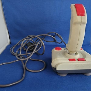 Peut inclure: Une manette de jeu joystick beige et grise vintage avec des boutons rouges et une poignée incurvée. La manette a un long câble gris et est conçue pour le jeu. Le joystick est sur une surface bleue.