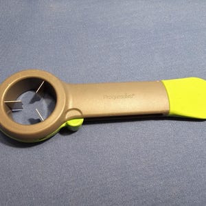 Puede incluir: Una herramienta de cocina gris y verde con la palabra "Progressive" impresa en el mango. La herramienta tiene un extremo circular con cuchillas y una punta de mango verde brillante. Está diseñada para abrir latas.