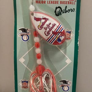 May include: A St. Louis Cardinals In-Line Spinner fishing lure on a green and white baseball-themed package. The lure features red, white, and blue accents, the team logo, and the Major League Baseball logo.