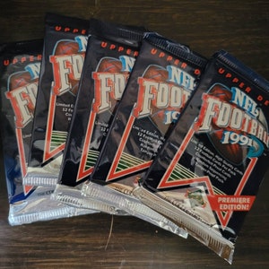 May include: Five unopened packs of 1991 Upper Deck NFL Football trading cards. The packs are silver and blue with the text "Upper Deck" and "NFL Football 1991" printed on them. The packs are arranged in a fan shape.