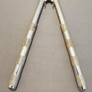 May include: A silver-colored nutcracker with textured handles. The nutcracker is open, displaying its jaws and the hinge mechanism. The handles have a cross-hatched pattern for grip, and the jaws have a serrated edge.