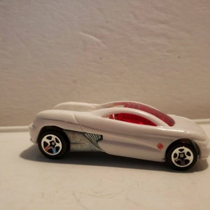 Peut inclure: Une voiture jouet blanche avec un pare-brise rouge et des roues noires. La voiture a un design élégant et aérodynamique et un petit emblème rouge sur le côté. Les roues sont noires avec des accents argentés.