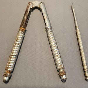 Puede incluir: Juego vintage de cascanueces y pincho plateados. El cascanueces tiene un diseño de tijera con mangos texturizados y un borde dentado. El pincho es delgado con una punta curva y un mango texturizado a juego. Ambos muestran signos de edad.