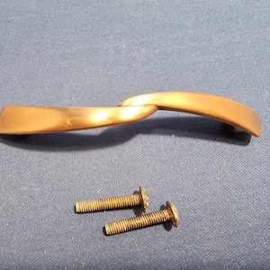 May include: A bronze-colored cabinet pull with a curved, twisted design. Two matching screws are included. The hardware is designed for easy installation and adds a decorative touch to furniture.