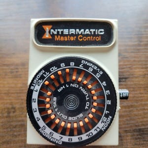 May include: A white Intermatic Master Control timer with a black dial and orange markings. The dial has numbers from 1 to 12 and the words "Morning", "Noon", "Evening", and "Midnite".