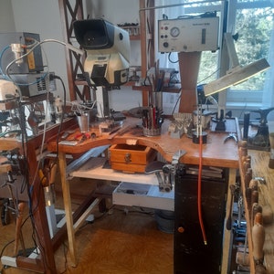 Peut inclure: Un établi avec une variété d'outils et d'équipements pour la fabrication de bijoux, notamment un microscope, une station de soudage et une unité Hydro-Star 2.0. L'établi est en bois et possède un tiroir en métal. Il y a également plusieurs outils et fournitures sur les étagères derrière l'établi.