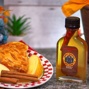 May include: A glass bottle of apple pie flavored honey with a cinnamon stick inside. The bottle has a brown label with the text "Honey Badgers Bee Farm" and a picture of three honey badgers. The bottle is sitting on a wooden surface next to a plate of apple pie with cinnamon sticks.