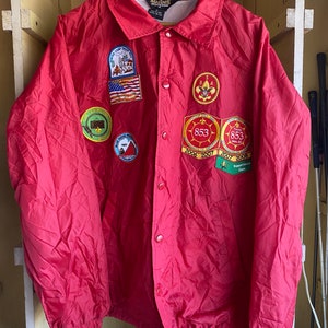 May include: Red nylon jacket with multiple embroidered patches, including a Boy Scouts of America patch, a "Troop 853" patch, and a "Pepperdine Cave" patch.