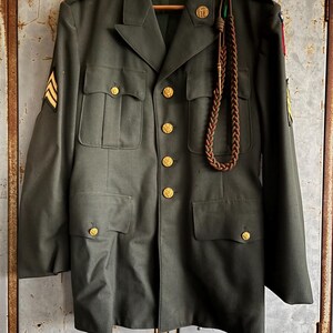 May include: Olive green military jacket with gold buttons and sergeant stripes on the left sleeve. The jacket has four front pockets and a decorative cord. The jacket is hanging on a wooden hanger.
