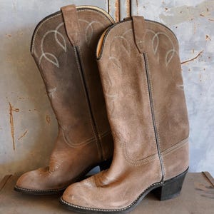 Könnte beinhalten: Ein Paar braune Wildleder-Cowbo Stiefel mit spitzer Zehenpartie und niedrigem Absatz. Die Stiefel haben ein dekoratives Steppmuster am Schaft und einen Reißverschluss an der Innenseite des Schafts.